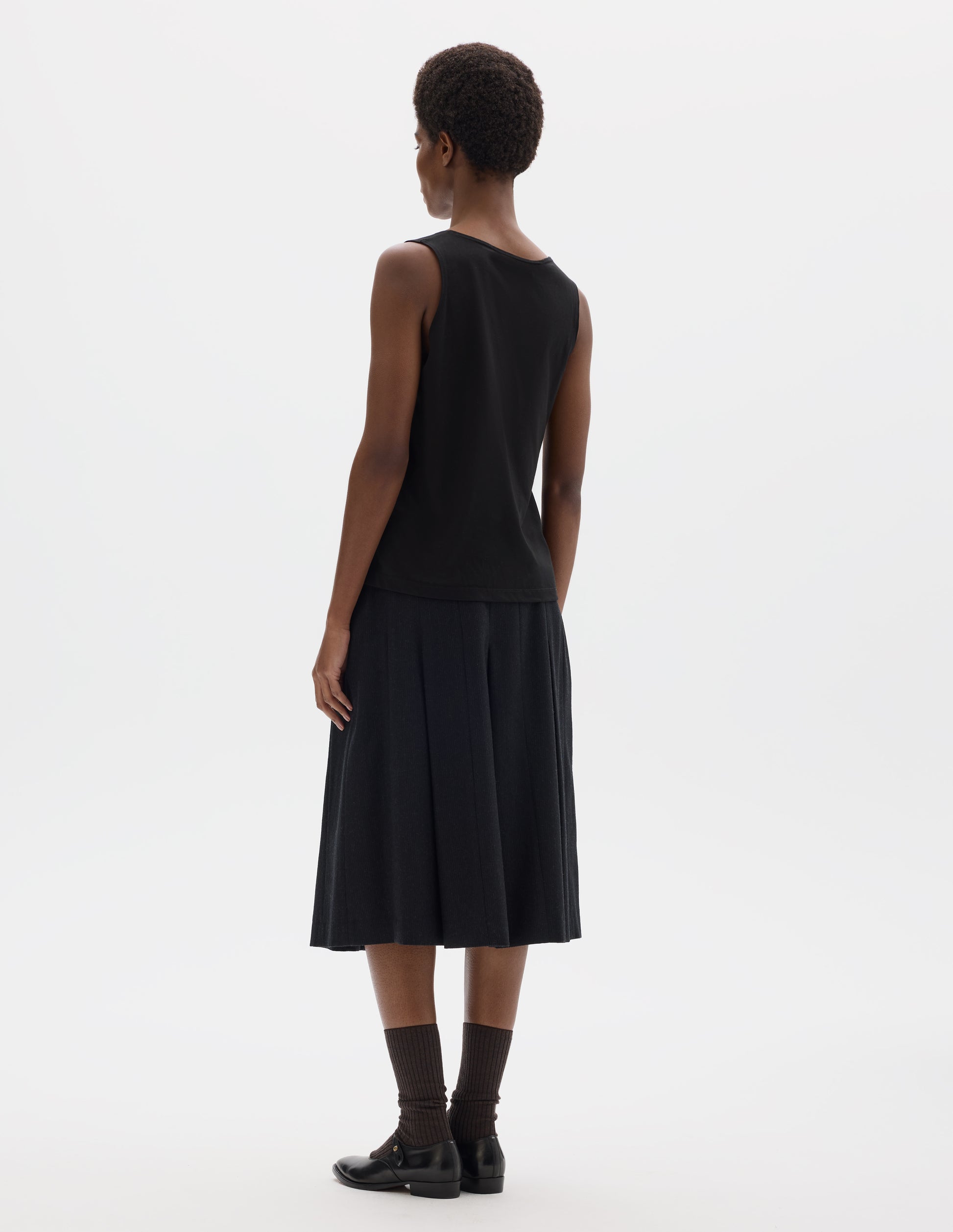 Full body image, back view of model wearing black sleeveless cotton vest.  Styled with dark grey skirt, dark brown socks and black leather shoes.