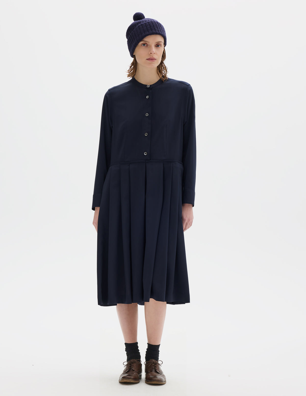 Full body image, front view of model wearing relaxed dark blue collarless wool midi dress with mother of pearl buttons on half placket, long sleeves with shirt cuffs, and pleated skirt with concealed side pockets. Styled with dark blue bobble hat, black socks and brown leather brogues.