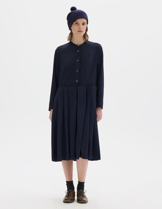 Full body image, front view of model wearing relaxed dark blue collarless wool midi dress with mother of pearl buttons on half placket, long sleeves with shirt cuffs, and pleated skirt with concealed side pockets. Styled with dark blue bobble hat, black socks and brown leather brogues.