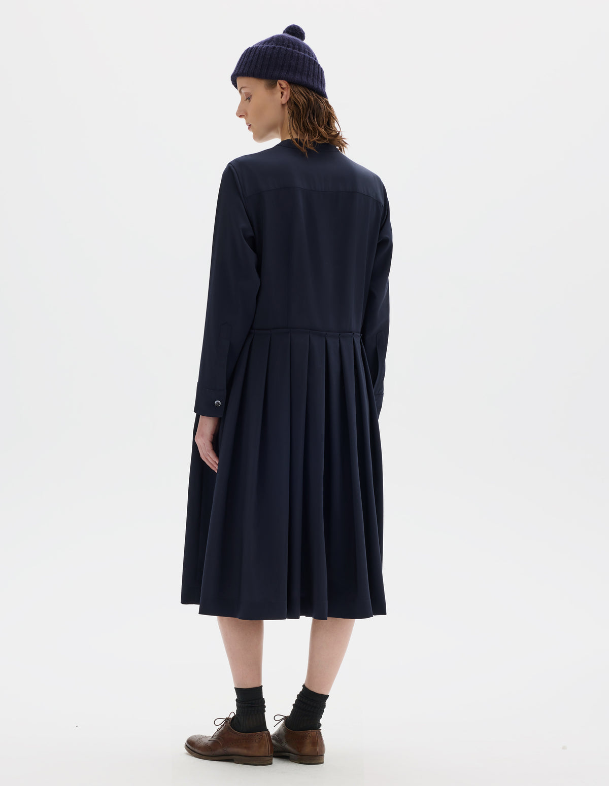 Full body image, back view of model wearing relaxed dark blue collarless wool midi dress with shirt yoke, long sleeves with mother of pearl buttons on cuffs and pleated skirt with concealed side pockets. Styled with dark blue bobble hat, black socks and brown leather brogues.