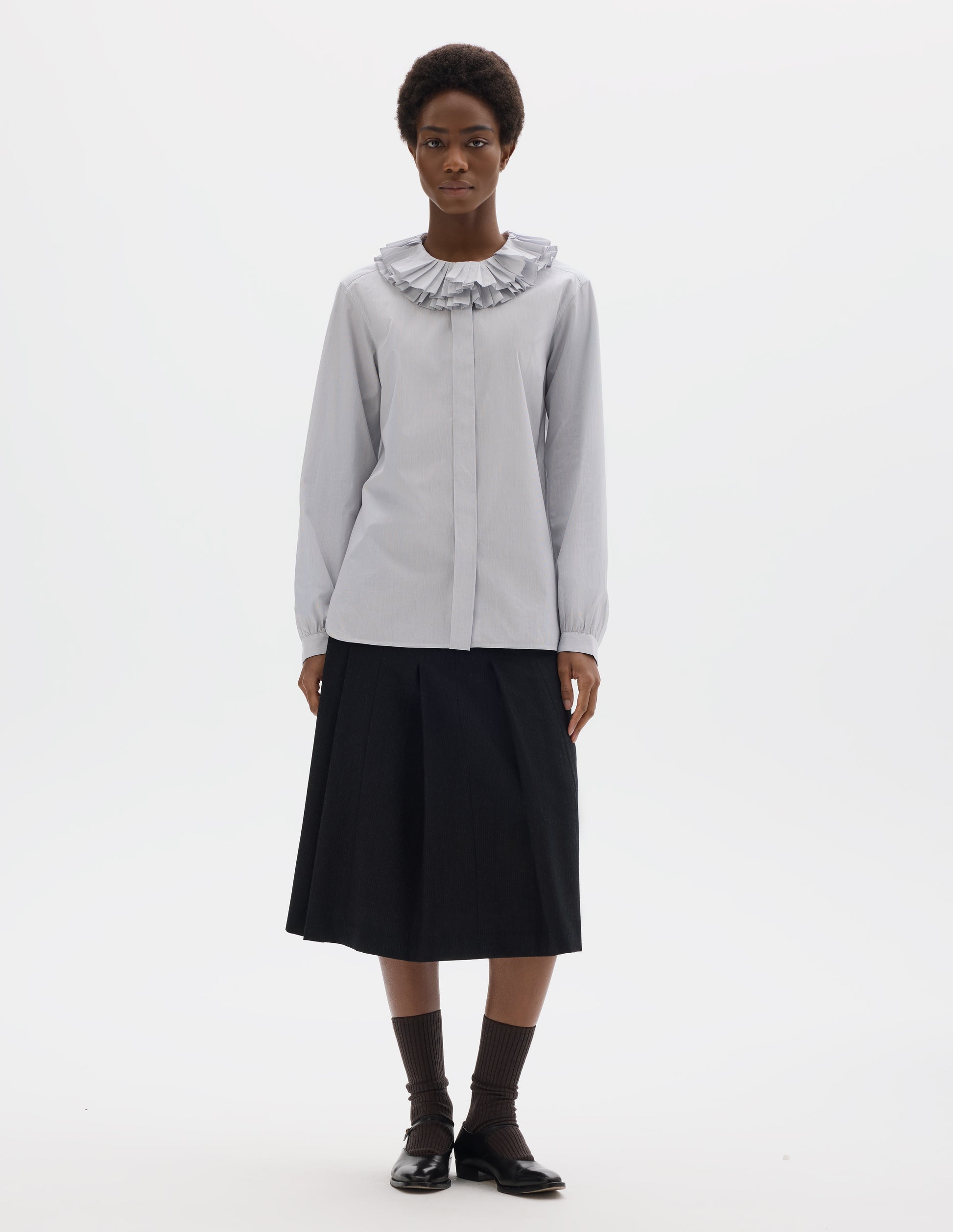 Full body image, front view of model wearing white cotton shirt with dark blue stripes, double ruffle collar, concealed placket with mother of pearl buttons, bracelet-length sleeves with mother of pearl buttons on narrow, gathered cuffs, and with curved hem. Styled with dark grey skirt, dark brown socks and black leather shoes.