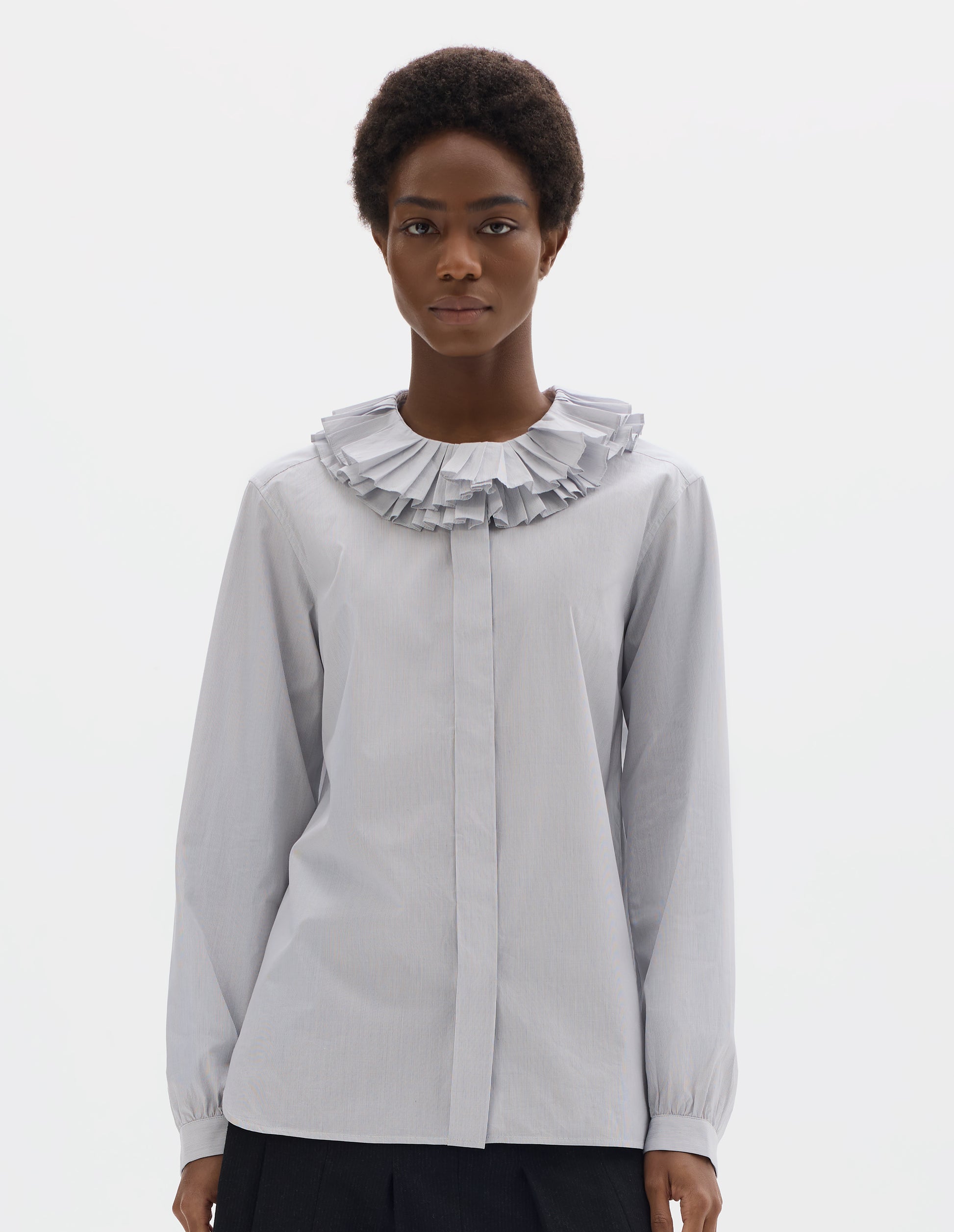 Cropped body image, front view of model wearing white cotton shirt with dark blue stripes, double ruffle collar, concealed placket with mother of pearl buttons, bracelet-length sleeves with mother of pearl buttons on narrow, gathered cuffs, and with curved hem. Styled with dark grey skirt.