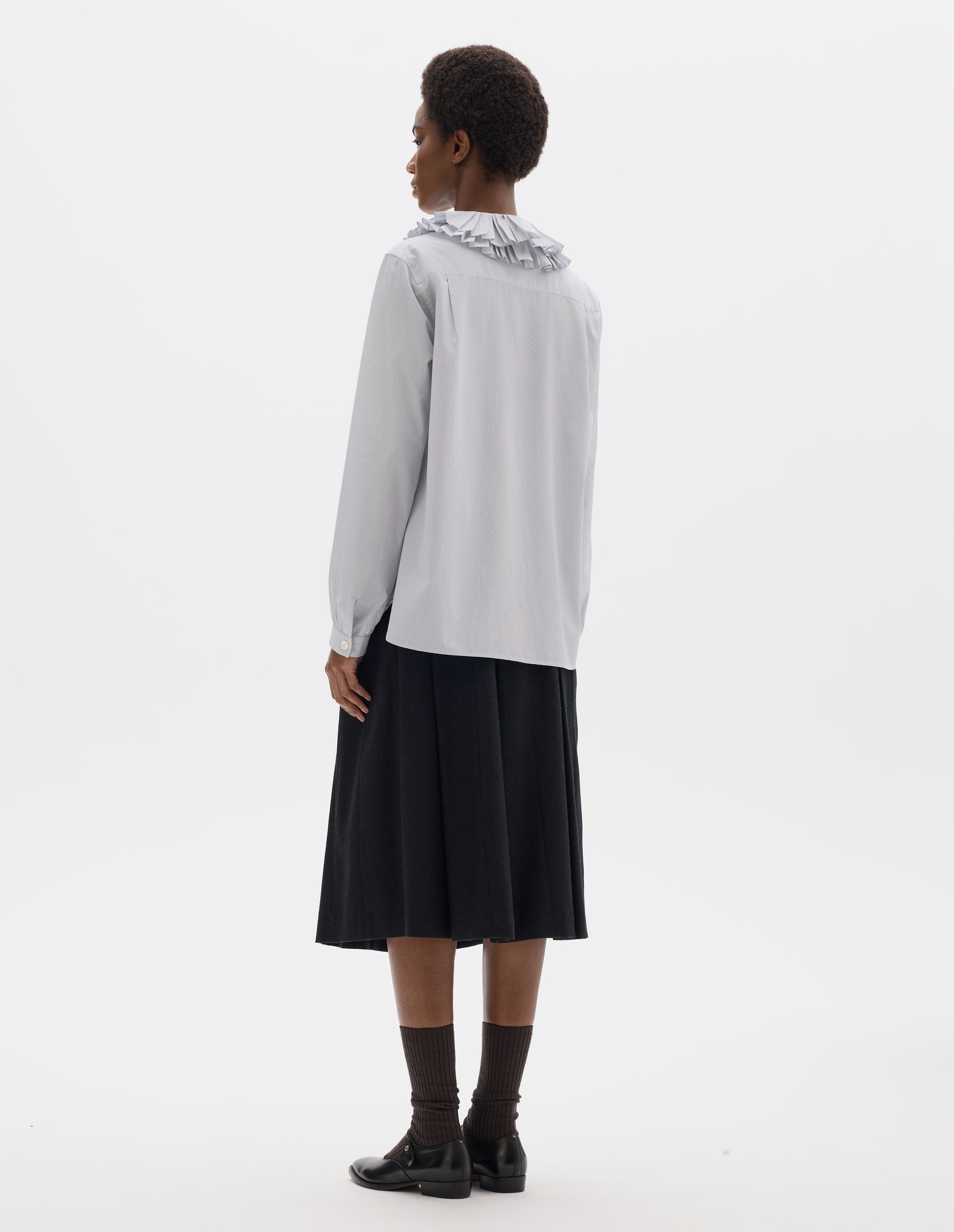 Full body image, back view of model wearing white cotton shirt with dark blue stripes, double ruffle collar, bracelet-length sleeves with mother of pearl buttons on narrow, gathered cuffs, and with curved hem. Styled with dark grey skirt, dark brown socks and black leather shoes.