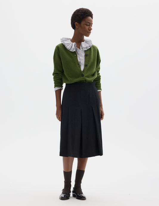 Full body image, front view of model wearing below knee length black wool skirt with dark grey stripes, pleats, narrow horn button-through waistband and side zip fly. Styled with white frilled shirt, green cardigan, dark brown socks and black leather shoes.
