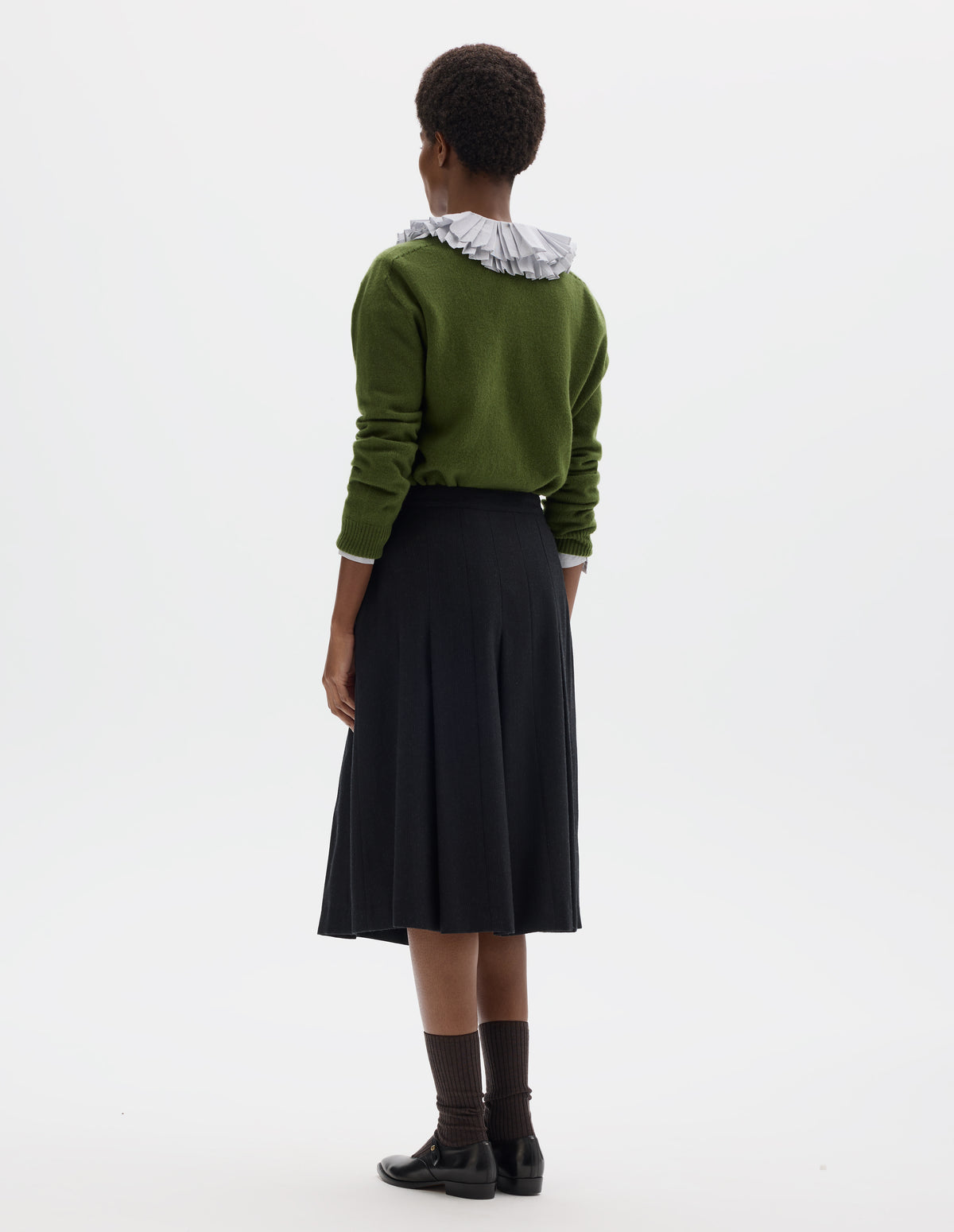 Full body image, back view of model wearing below knee length black wool skirt with dark grey stripes, pleats, narrow horn button-through waistband and side zip fly. Styled with white frilled shirt, green cardigan, dark brown socks and black leather shoes.
