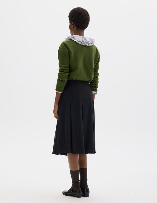 Full body image, back view of model wearing below knee length black wool skirt with dark grey stripes, pleats, narrow horn button-through waistband and side zip fly. Styled with white frilled shirt, green cardigan, dark brown socks and black leather shoes.