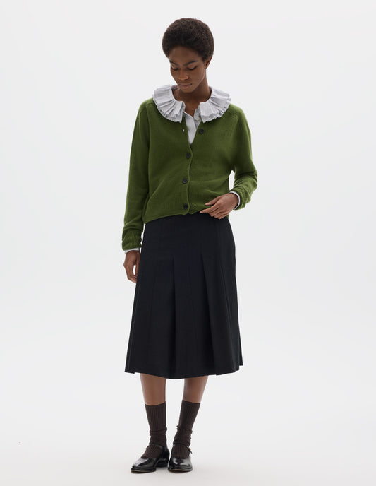 Full body image, front view of model wearing green merino cashmere fully fashioned cardigan with horn buttons, and with ribbed crew neck, cuffs and hem. Styled over white ruffle shirt, with black skirt, dark brown socks and black leather shoes.