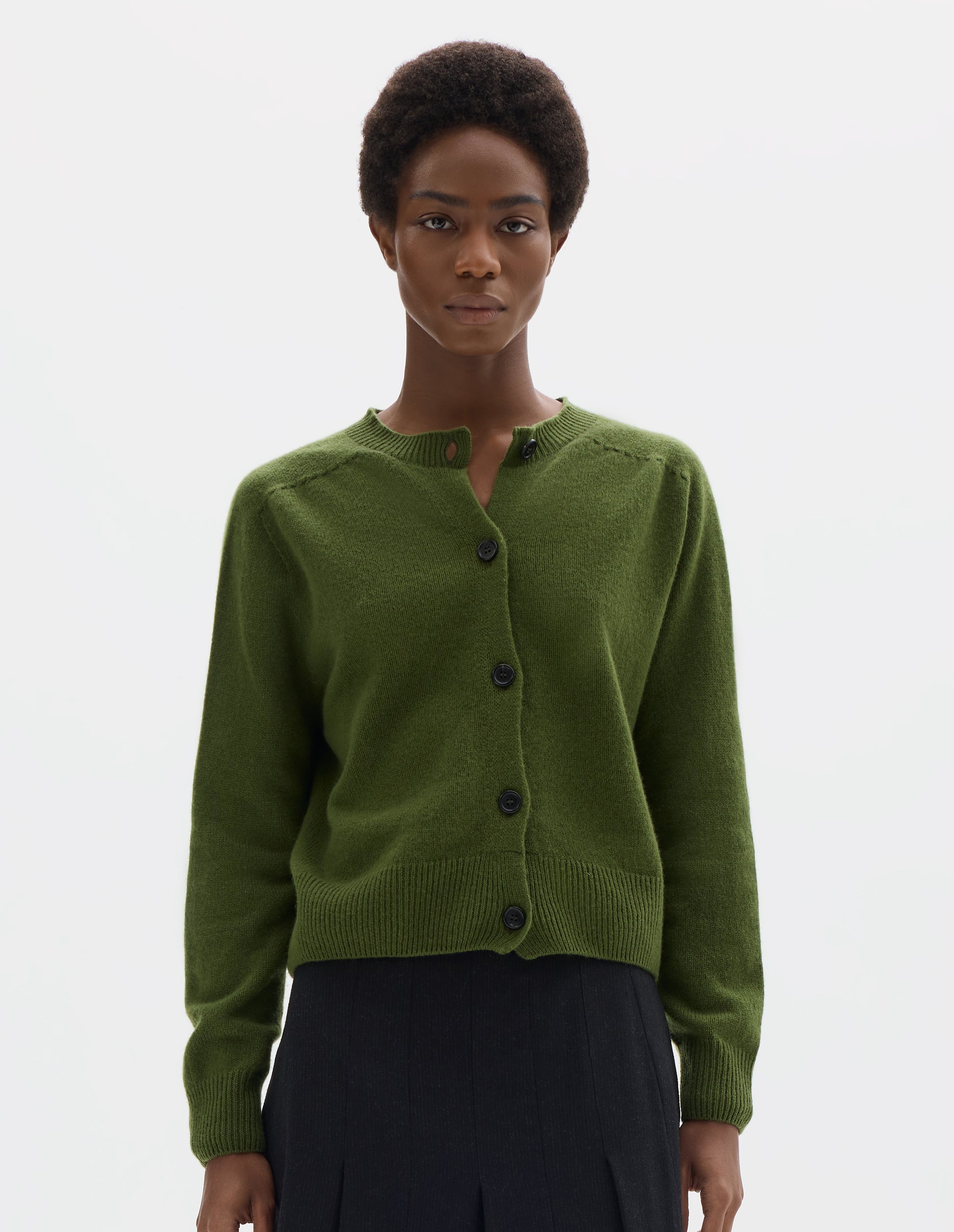 Cropped body image, front view of model wearing green merino cashmere fully fashioned cardigan with horn buttons, and with ribbed crew neck, cuffs and hem. Styled with black skirt.