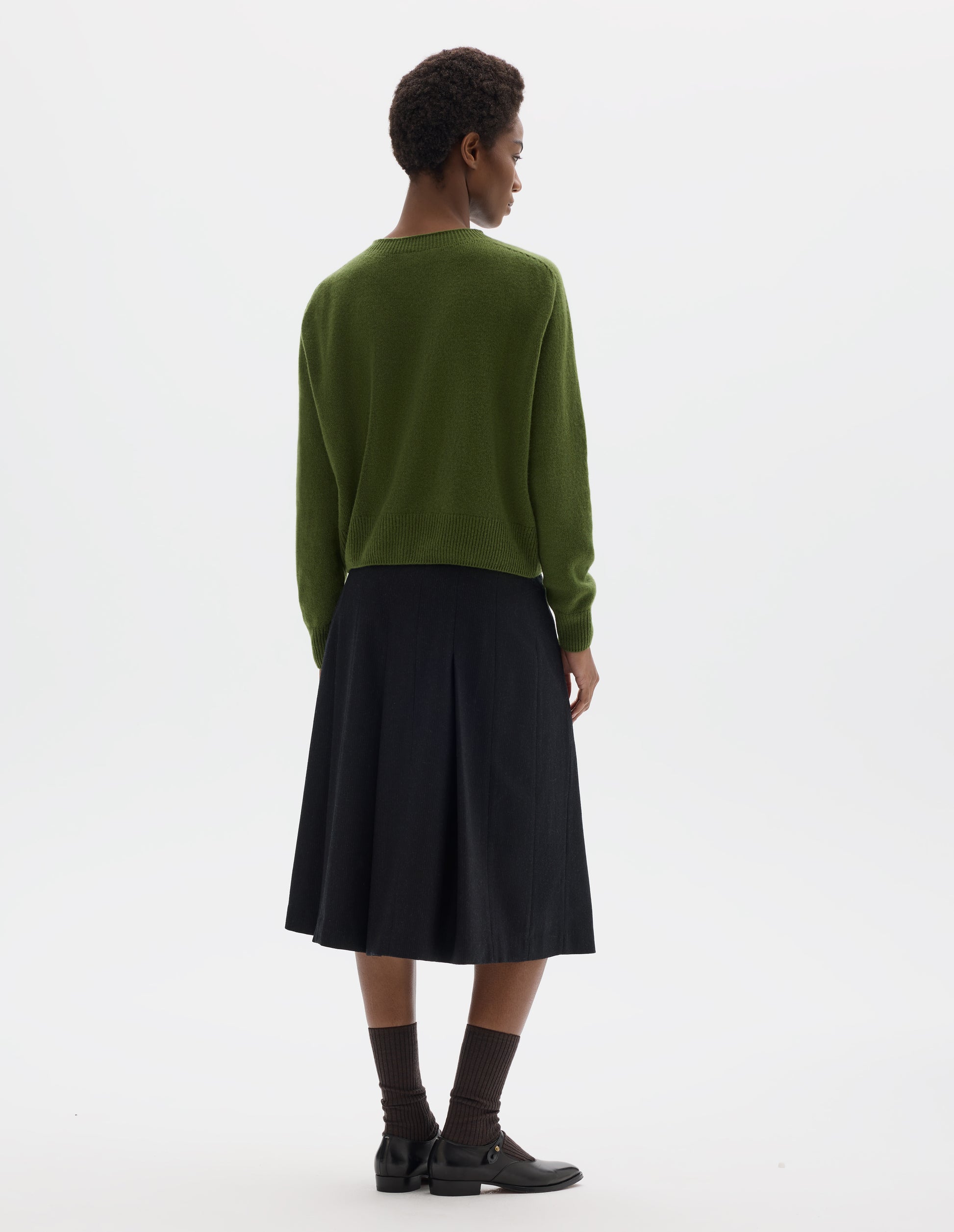 Full body image, back view of model wearing green merino cashmere fully fashioned cardigan with ribbed crew neck, cuffs and hem. Styled with black skirt, dark brown socks and black leather shoes.