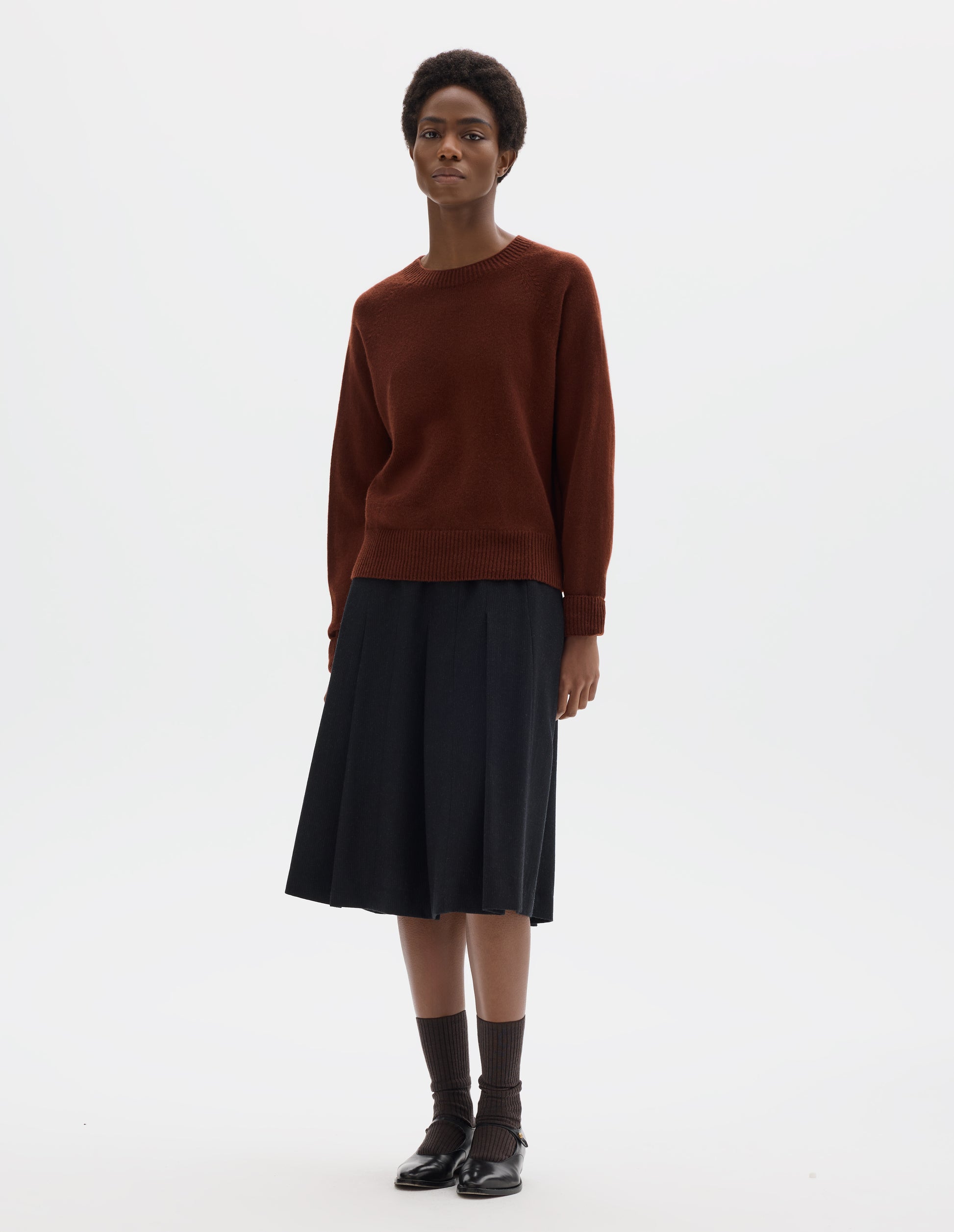 Full body image, front view of model wearing brown fully fashioned seamless merino cashmere jumper with crew neck, raglan sleeves, and with ribbed neck, cuffs and hem. Styled with dark grey skirt, brown socks and black leather shoes.