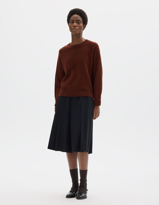 Full body image, front view of model wearing brown fully fashioned seamless merino cashmere jumper with crew neck, raglan sleeves, and with ribbed neck, cuffs and hem. Styled with dark grey skirt, brown socks and black leather shoes.