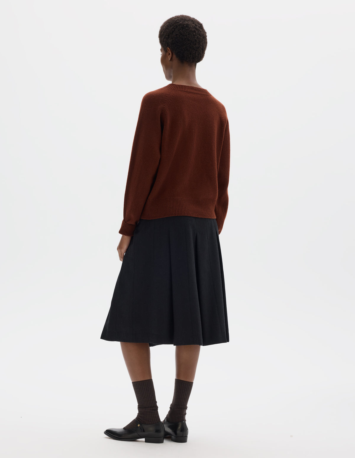 Full body image, back view of model wearing brown fully fashioned seamless merino cashmere jumper with crew neck, raglan sleeves, and with ribbed neck, cuffs and hem. Styled with dark grey skirt, brown socks and black leather shoes.