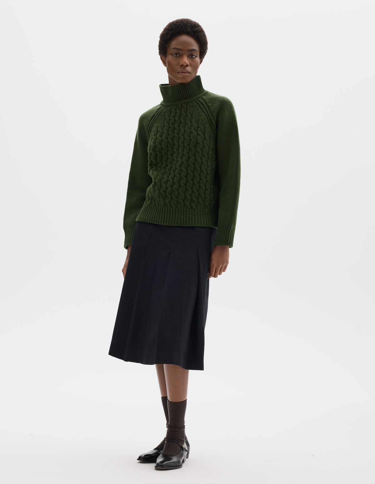 Full body image, front view of model wearing relaxed green merino jumper with ribbed high neck collar, front cable design, raglan sleeves with ribbed side seams, cuffs and hem. Styled with black skirt, dark brown socks and black leather shoes.