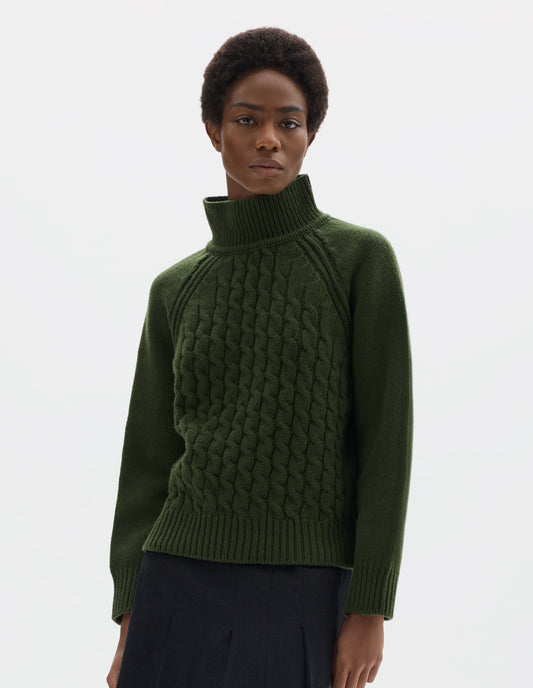 Cropped body image, front view of model wearing relaxed green merino jumper with ribbed high neck collar, front cable design, raglan sleeves with ribbed side seams, cuffs and hem. Styled with black skirt.