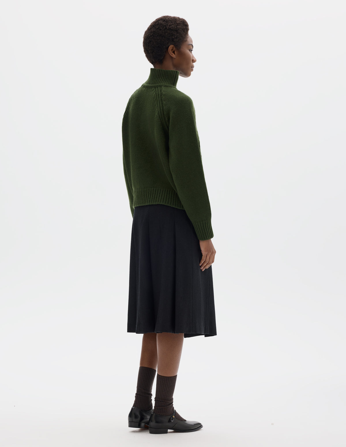 Full body image, back view of model wearing relaxed green merino jumper with plain knitted back, raglan sleeves with ribbed side seams and ribbed high neck collar, cuffs and hem. Styled with black skirt, dark brown socks and black leather shoes.