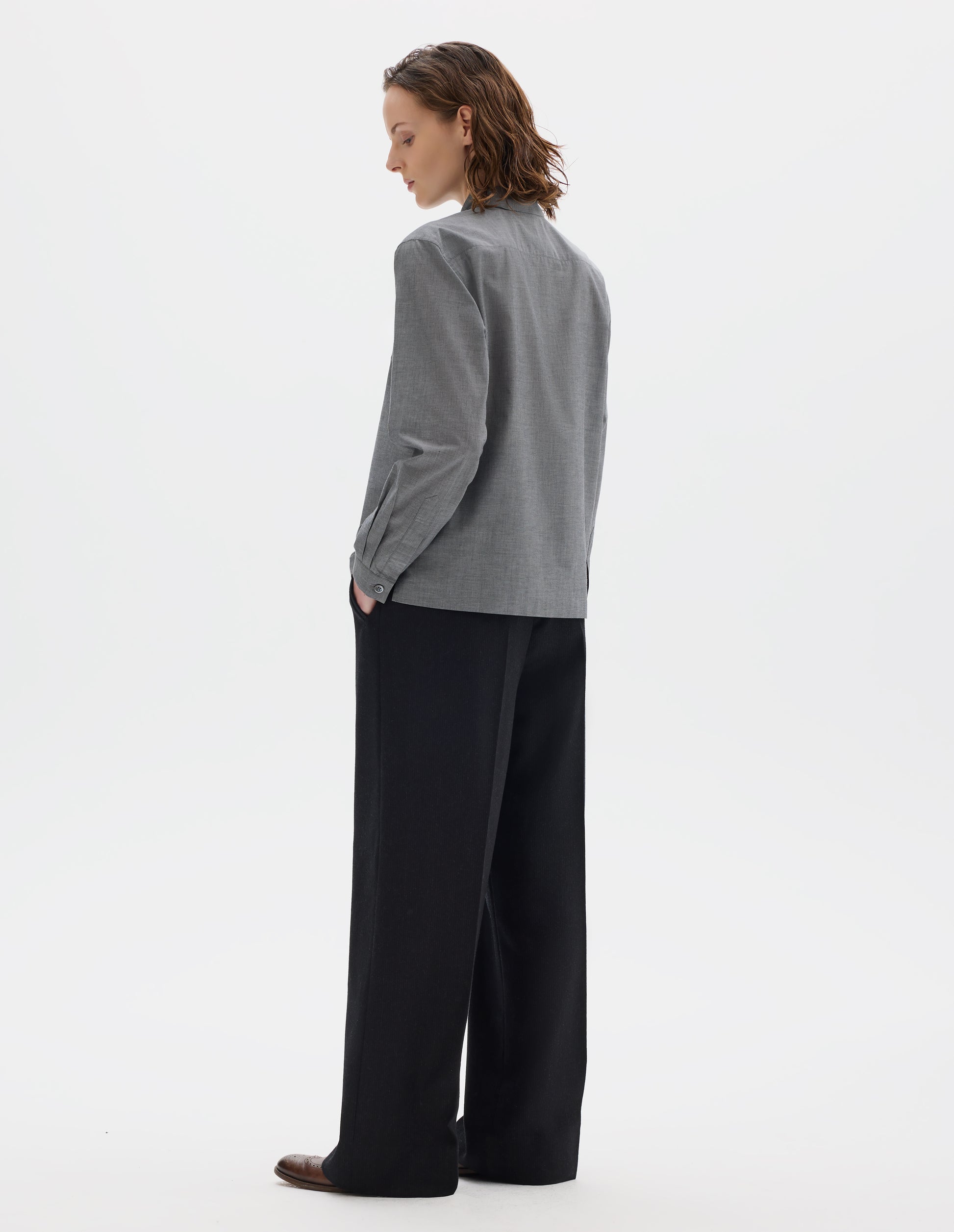 Full body image, back view of model wearing grey cotton shirt with mother of pearl buttons on narrow cuffs with pleats, and with square side split hem. Styled with black trousers and brown leather brogues.