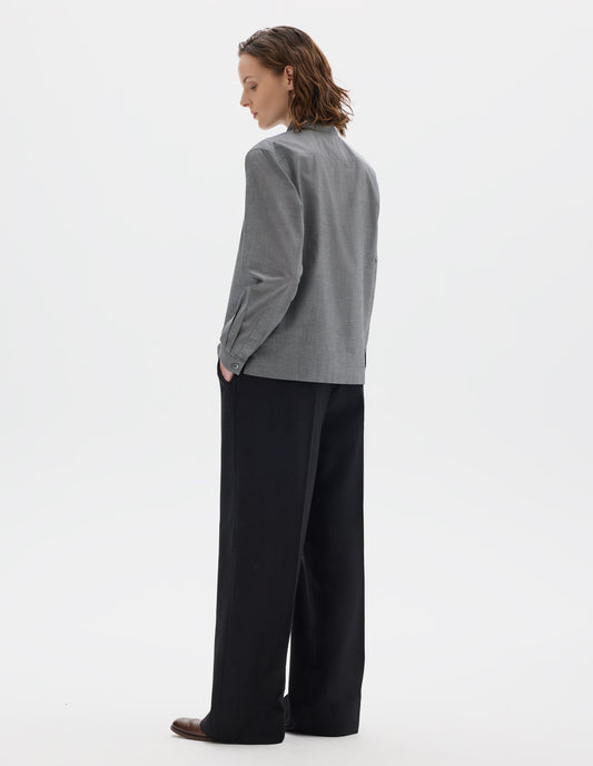 Full body image, back view of model wearing grey cotton shirt with mother of pearl buttons on narrow cuffs with pleats, and with square side split hem. Styled with black trousers and brown leather brogues.