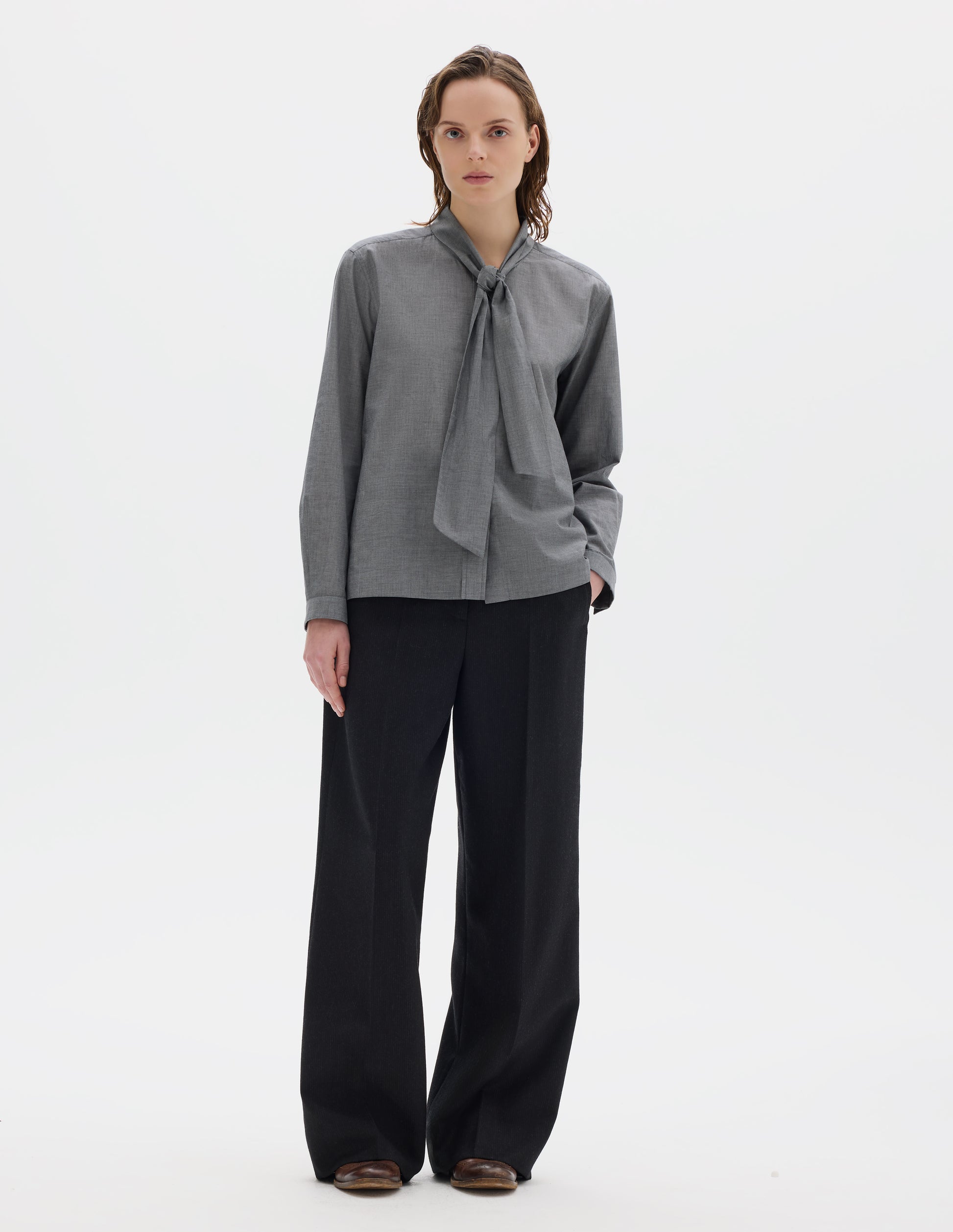 Full body image, front view of model wearing grey cotton shirt with attached loose tie, lipped placket, narrow cuffs and with square side split hem. Styled with black trousers and brown leather brogues.
