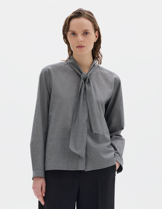 Cropped body image, front view of model wearing grey cotton shirt with attached loose tie, lipped placket, narrow cuffs and with square side split hem. Styled with black trousers.