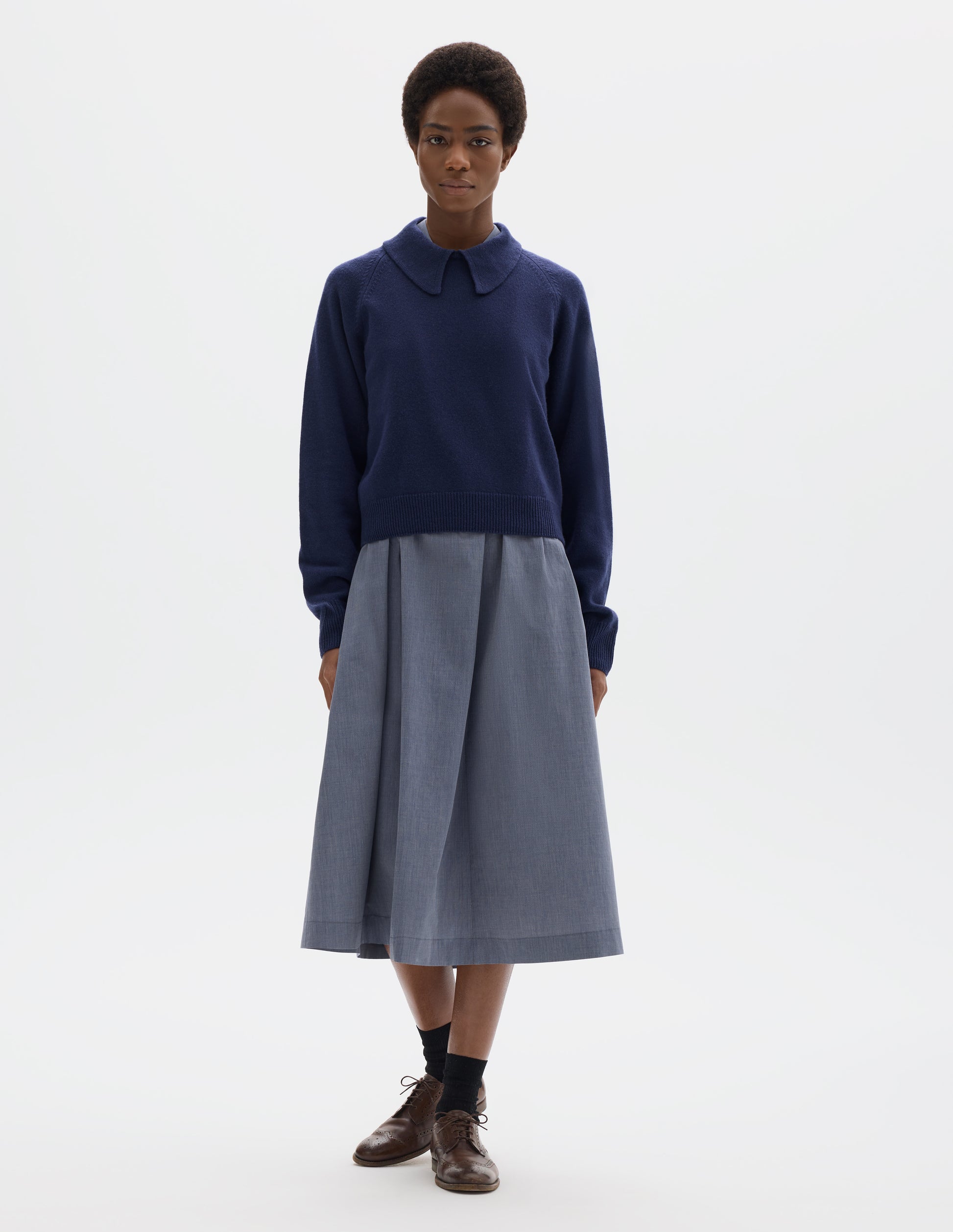 Full body image, front view of model wearing dark blue cropped merino jumper with exaggerated collar, raglan sleeves, and with ribbed cuffs and hem. Styled over blue midi dress, and with black socks and brown leather brogues.