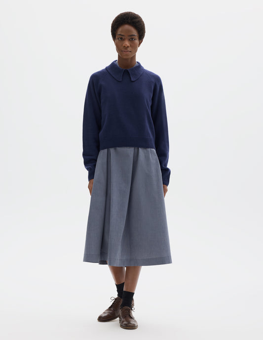 Full body image, front view of model wearing dark blue cropped merino jumper with exaggerated collar, raglan sleeves, and with ribbed cuffs and hem. Styled over blue midi dress, and with black socks and brown leather brogues.