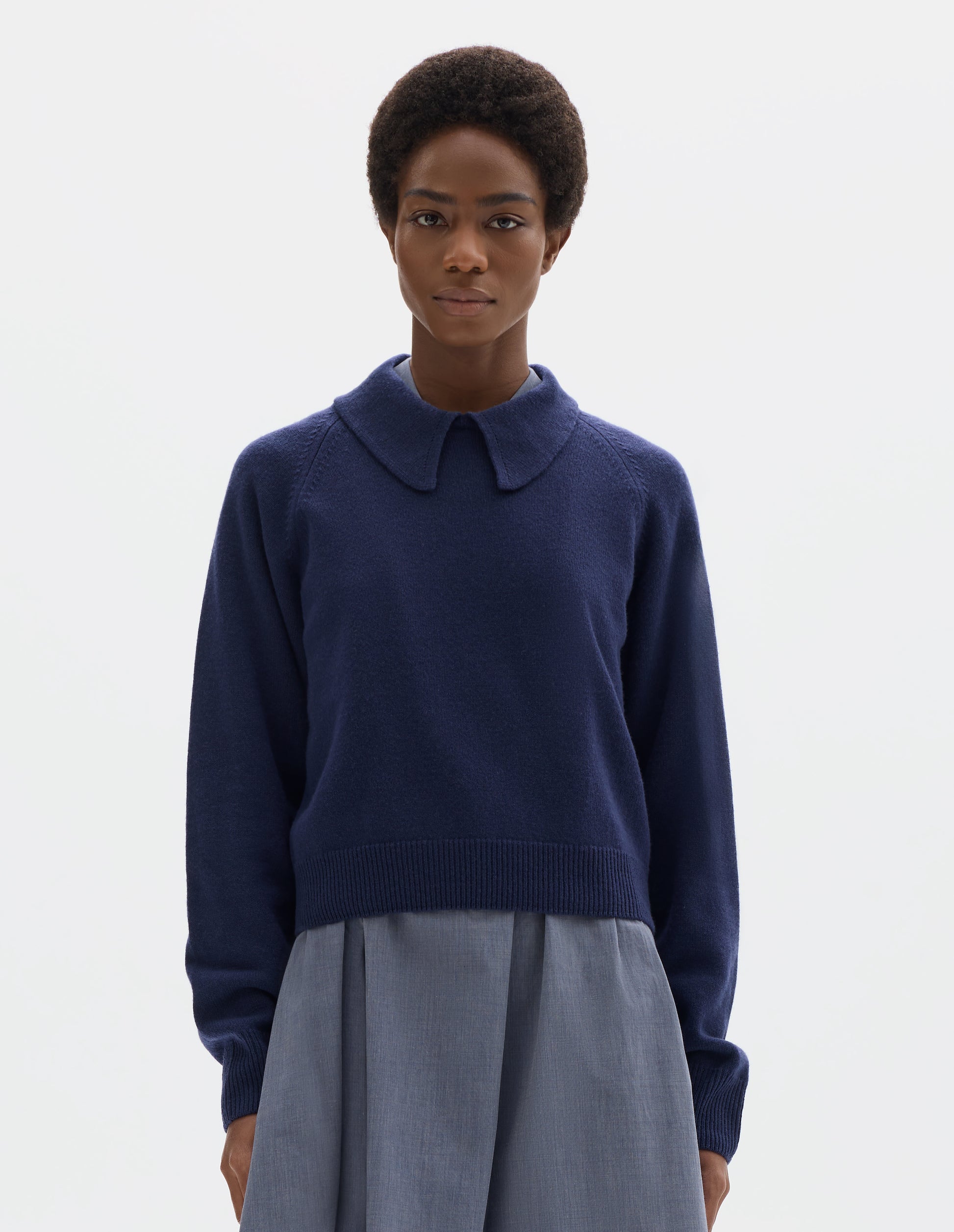 Cropped body image, front view of model wearing dark blue cropped merino jumper with exaggerated collar, raglan sleeves, and with ribbed cuffs and hem. Styled over blue midi dress.