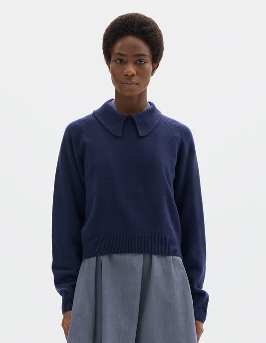 Cropped body image, front view of model wearing dark blue cropped merino jumper with exaggerated collar, raglan sleeves, and with ribbed cuffs and hem. Styled over blue midi dress.