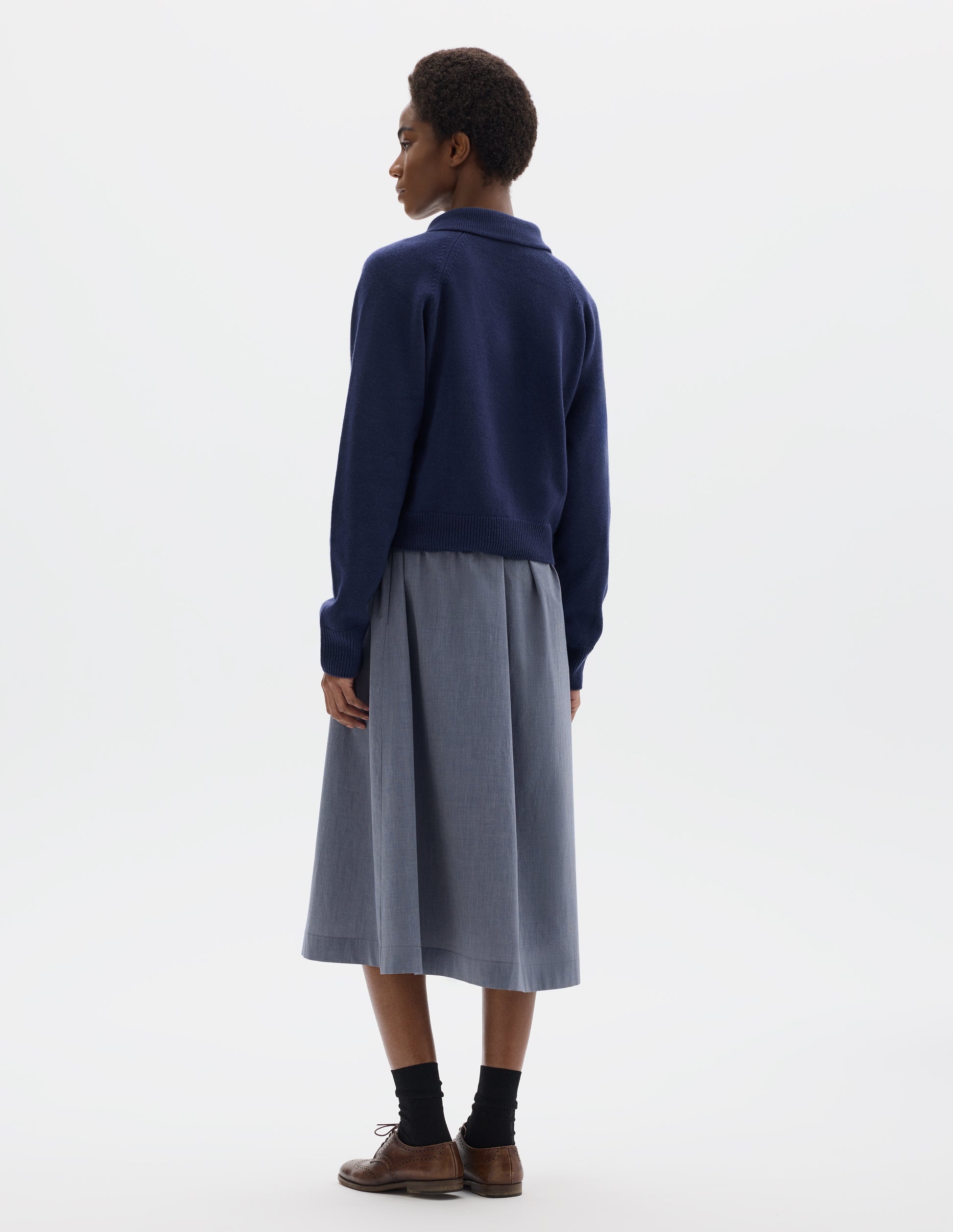 Full body image, back view of model wearing dark blue cropped merino jumper with collar, raglan sleeves, and with ribbed cuffs and hem. Styled over blue midi dress, and with black socks and brown leather brogues.