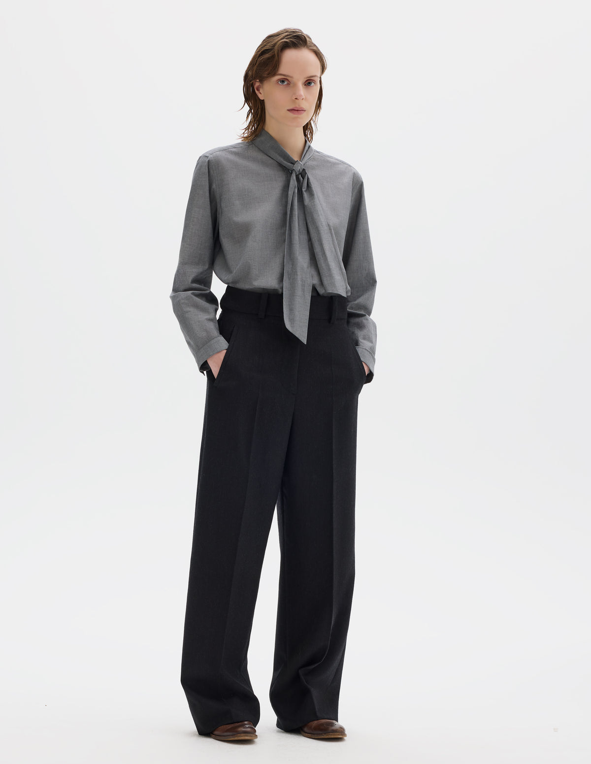 Full body image, front view of model wearing black high waisted wool trousers with dark grey stripes, belt loops on wide waistband, zip fly and horn button fastening, side adjusters, slanted front welt pockets, and wide leg. Styled with grey shirt and brown leather brogues.