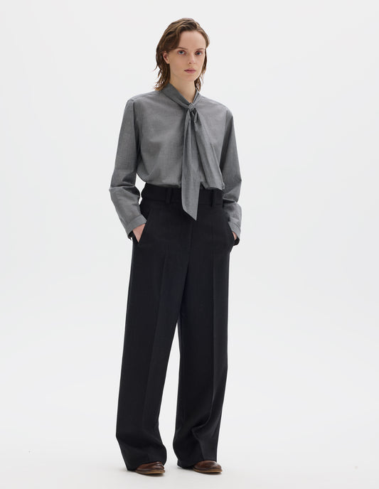 Full body image, front view of model wearing black high waisted wool trousers with dark grey stripes, belt loops on wide waistband, zip fly and horn button fastening, side adjusters, slanted front welt pockets, and wide leg. Styled with grey shirt and brown leather brogues.
