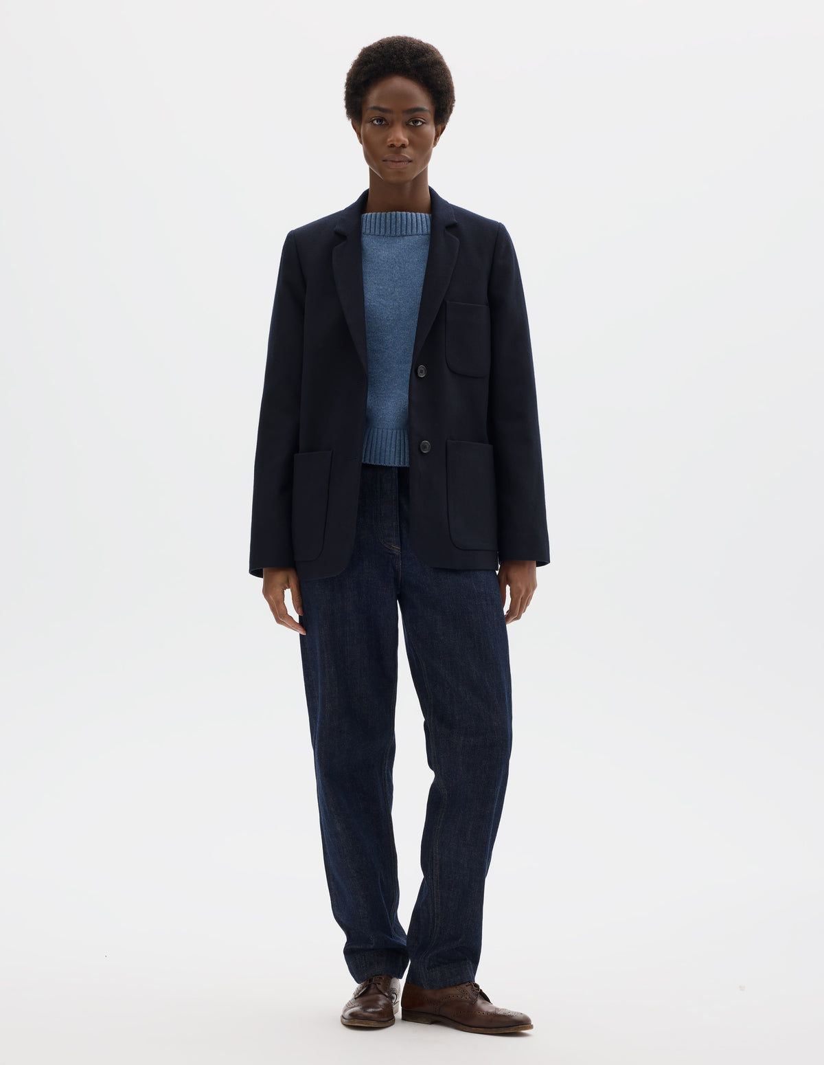 Full body image, front view of model wearing relaxed dark blue wool blazer with narrow collar and horn button-through revere, three bluff patch pockets, rounded details on hem and pockets. Styled over light blue knitted jumper, dark blue jeans and brown leather brogues.