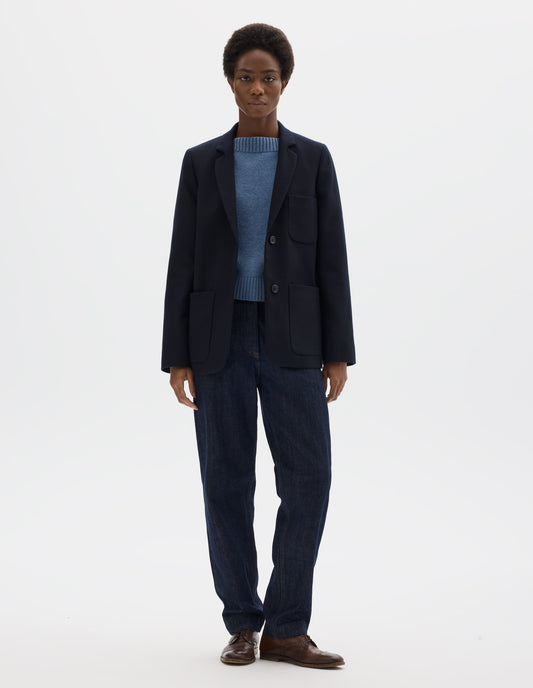 Full body image, front view of model wearing relaxed dark blue wool blazer with narrow collar and horn button-through revere, three bluff patch pockets, rounded details on hem and pockets. Styled over light blue knitted jumper, dark blue jeans and brown leather brogues.