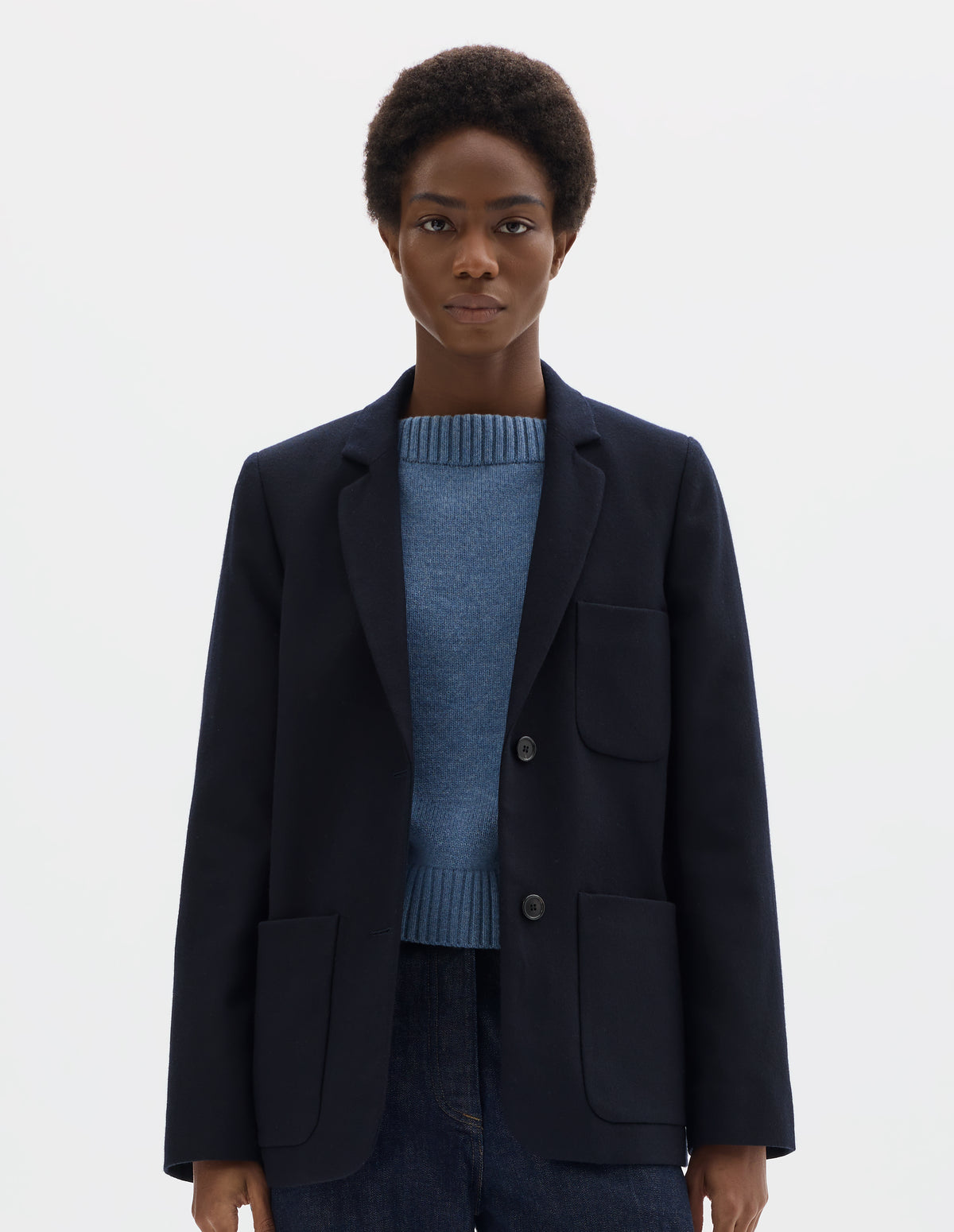 Cropped body image, front view of model wearing relaxed dark blue wool blazer with narrow collar and horn button-through revere, three bluff patch pockets, rounded details on hem and pockets. Styled with buttons undone over light blue knitted jumper  and dark blue jeans.