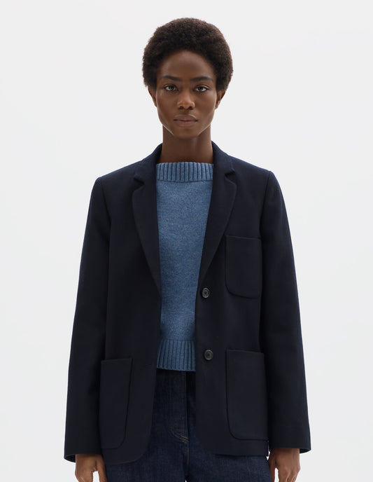 Cropped body image, front view of model wearing relaxed dark blue wool blazer with narrow collar and horn button-through revere, three bluff patch pockets, rounded details on hem and pockets. Styled with buttons undone over light blue knitted jumper  and dark blue jeans.