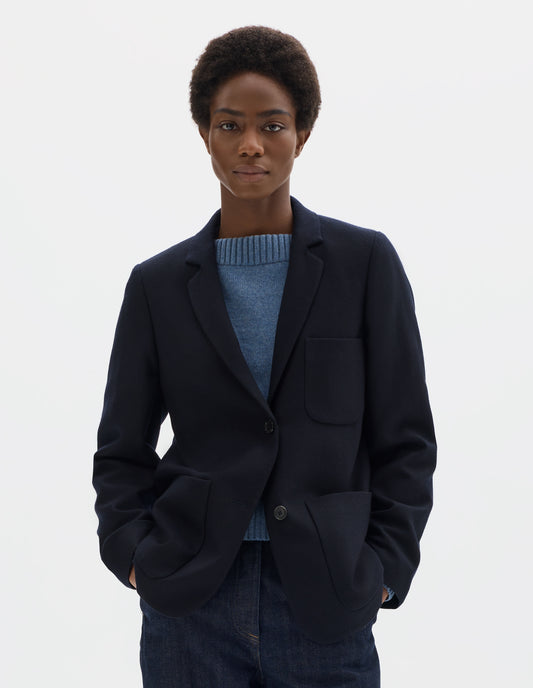 Cropped body image, front view of model wearing relaxed dark blue wool blazer with narrow collar and horn button-through revere, three bluff patch pockets, rounded details on hem and pockets. Styled with top button done up over light blue knitted jumper with dark blue jeans.
