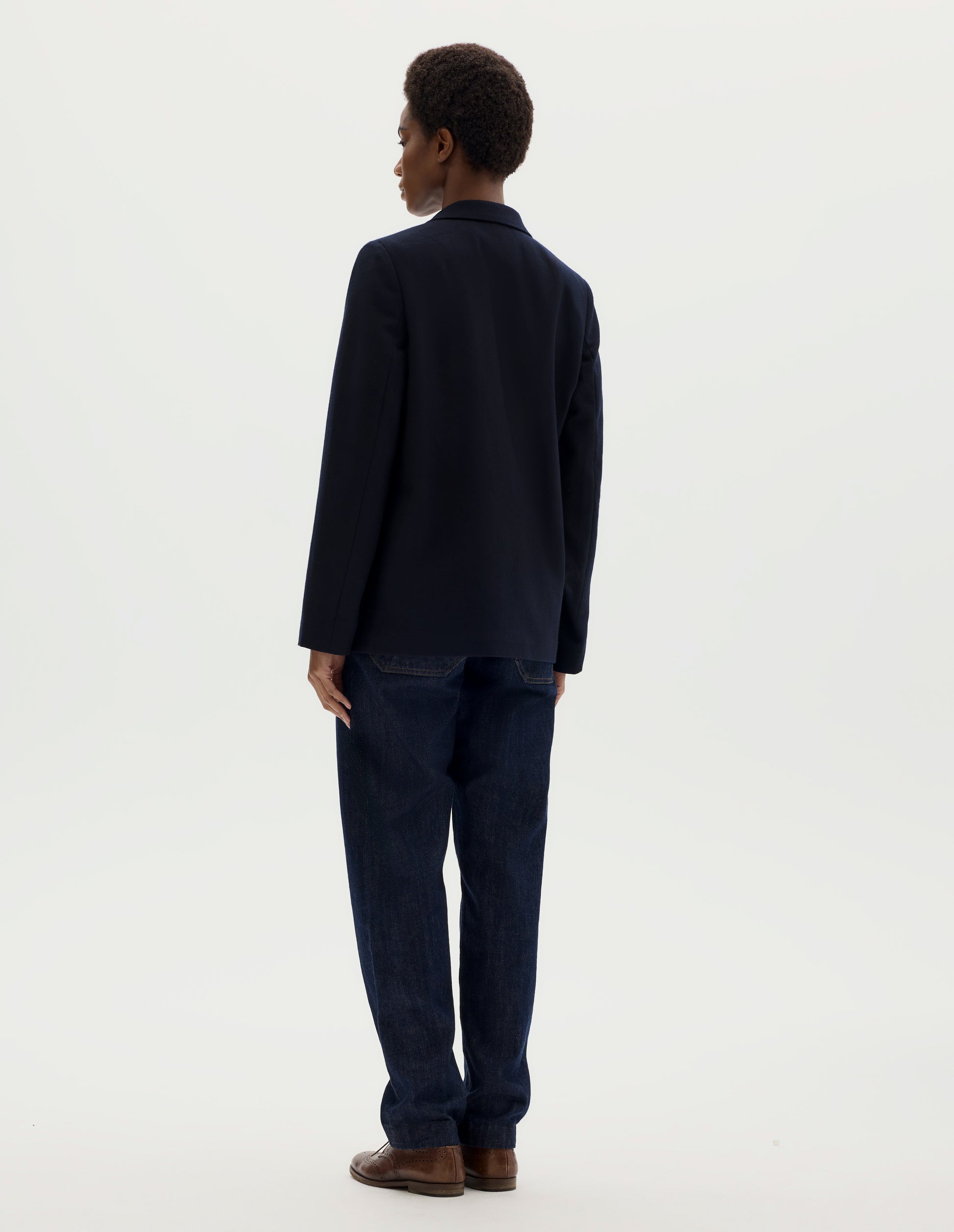 Full body image, back view of model wearing relaxed dark blue wool blazer with narrow collar. Styled with dark blue jeans and brown leather brogues.