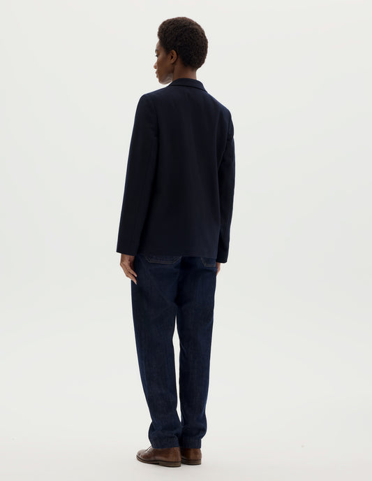 Full body image, back view of model wearing relaxed dark blue wool blazer with narrow collar. Styled with dark blue jeans and brown leather brogues.