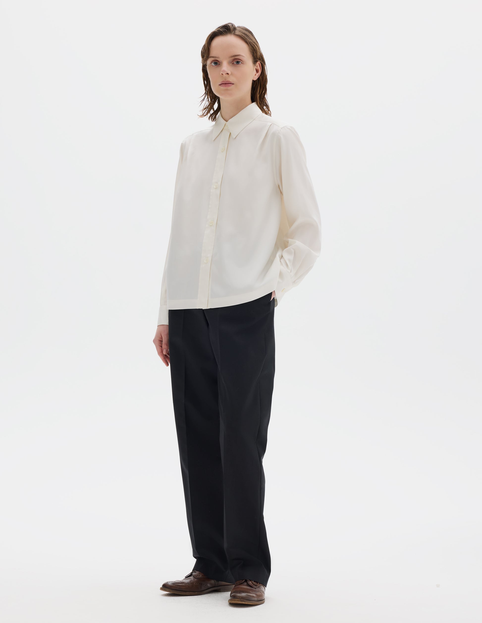 Full body image, front view of model wearing white silk shirt with mother of pearl buttons on placket and angled cuffs, narrow shoulder yoke with two pleats, and with vent split hem. Styled with black trousers and brown leather brogues.