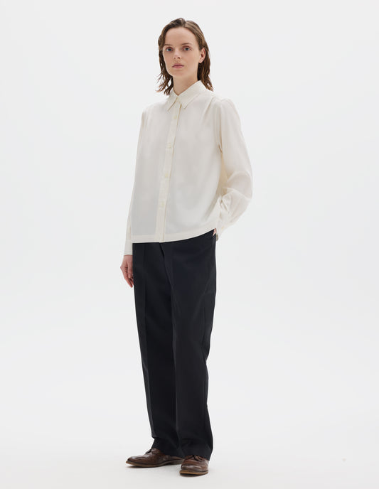 Full body image, front view of model wearing white silk shirt with mother of pearl buttons on placket and angled cuffs, narrow shoulder yoke with two pleats, and with vent split hem. Styled with black trousers and brown leather brogues.