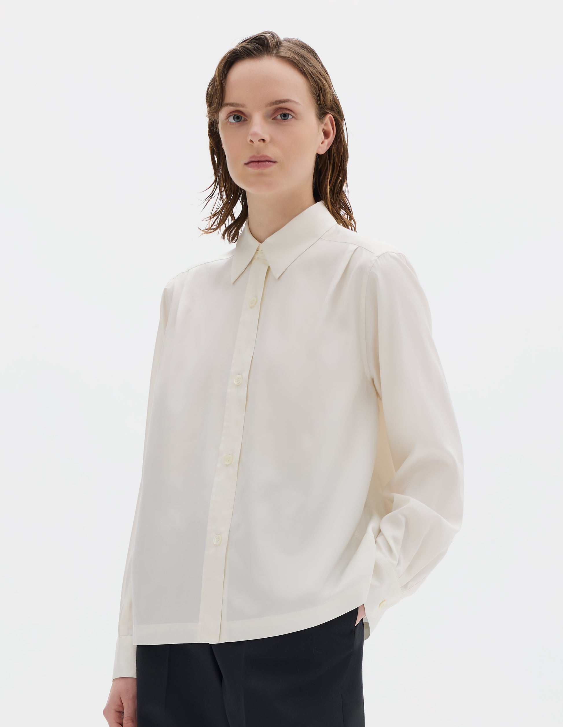 Cropped body image, front view of model wearing white silk shirt with mother of pearl buttons on placket and angled cuffs, narrow shoulder yoke with two pleats, and with vent split hem. Styled with black trousers.