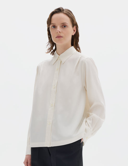 Cropped body image, front view of model wearing white silk shirt with mother of pearl buttons on placket and angled cuffs, narrow shoulder yoke with two pleats, and with vent split hem. Styled with black trousers.