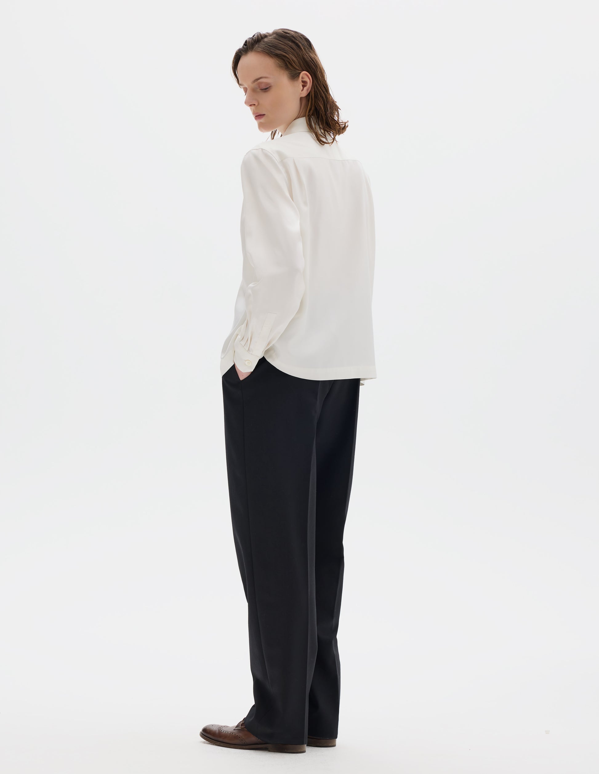 Full body image, back view of model wearing white silk shirt with narrow shoulder yoke with two pleats, mother of pearl buttons on angled cuffs and with vent split hem. Styled with black trousers and brown leather brogues.