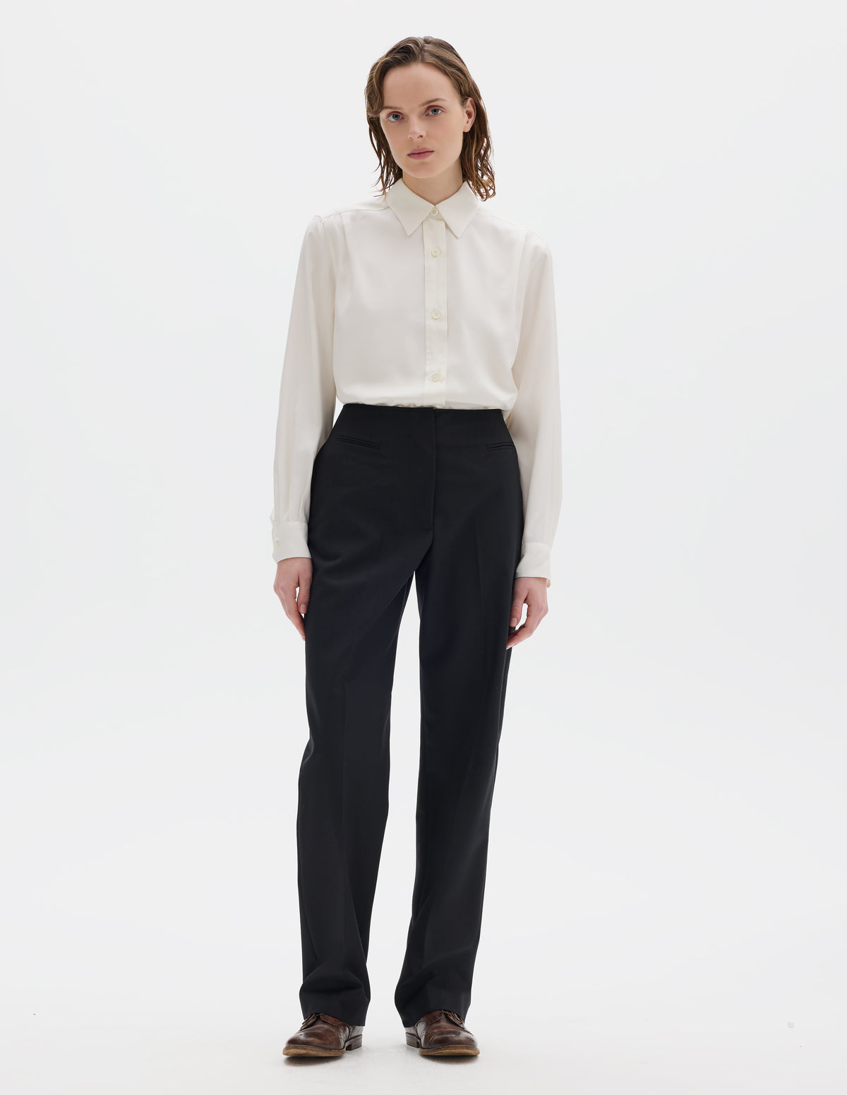 Full body image, front view of model wearing black tapered wool trousers with cotton fine-binding waistband, zip fly with hook and bar fastening, small front welt pockets and side pockets. Styled with beige shirt and brown leather brogues.