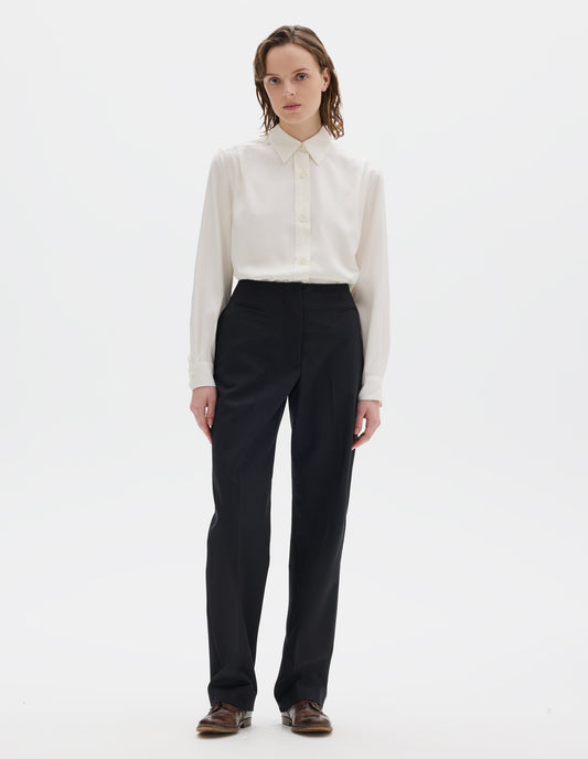 Full body image, front view of model wearing black tapered wool trousers with cotton fine-binding waistband, zip fly with hook and bar fastening, small front welt pockets and side pockets. Styled with beige shirt and brown leather brogues.