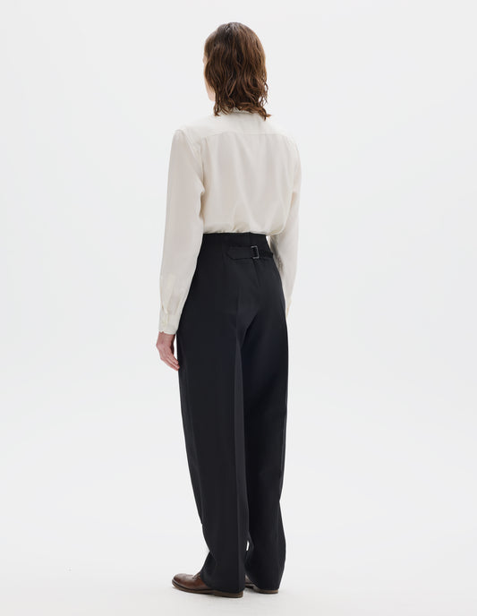 Full body image, back view of model wearing black tapered wool trousers with cotton fine-binding waistband and cinch detail on back. Styled with beige shirt and brown leather brogues.
