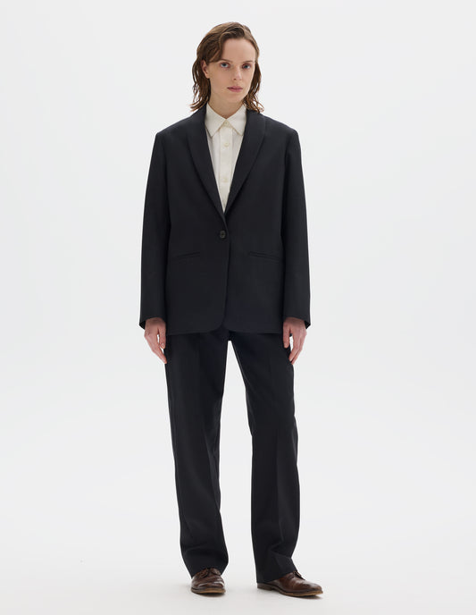 Full body image, front view of model wearing relaxed black wool blazer with same fabric lapel, single horn button fastening, horn button-through cuffs and slanted welt pockets. Styled over beige shirt, and with matching trousers and brown leather brogues.