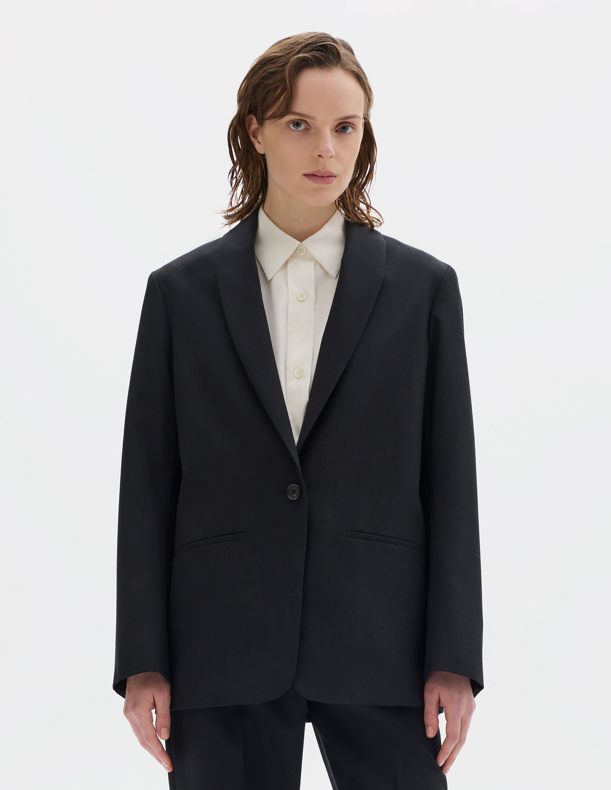 Cropped body image, front view of model wearing relaxed black wool blazer with same fabric lapel, single horn button fastening, horn button-through cuffs and slanted welt pockets. Styled over beige shirt, and with matching trousers.