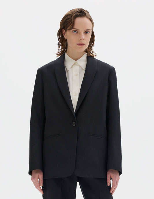 Cropped body image, front view of model wearing relaxed black wool blazer with same fabric lapel, single horn button fastening, horn button-through cuffs and slanted welt pockets. Styled over beige shirt, and with matching trousers.