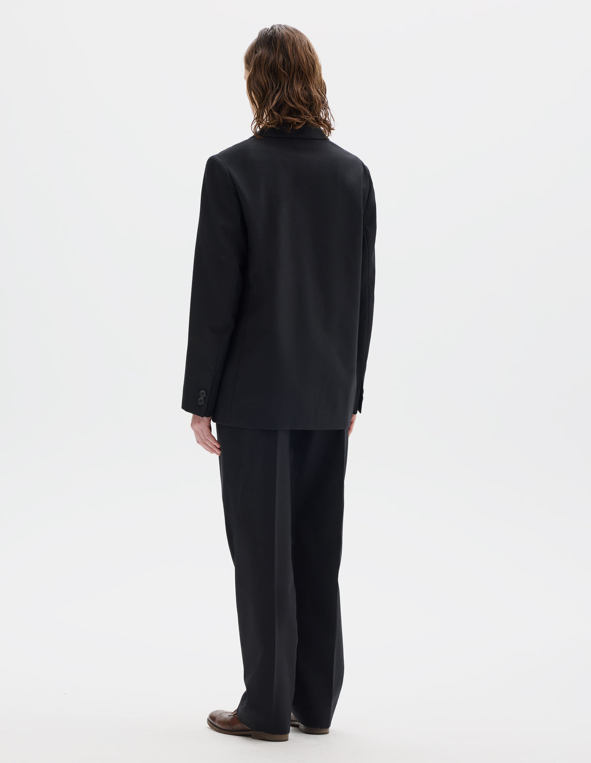 Full body image, back view of model wearing black relaxed wool blazer with horn buttons on cuffs. Styled with matching trousers and brown leather brogues.