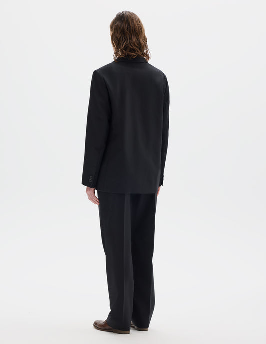 Full body image, back view of model wearing black relaxed wool blazer with horn buttons on cuffs. Styled with matching trousers and brown leather brogues.
